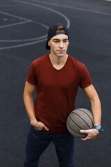 Handsome cool student guy with a cap in a T-shirt holds a basketball and stands at the stadium
