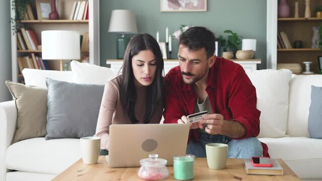 Smiling young couple making shopping online with credit card and laptop at home. Two happy people holding debit card while buying on e-commerce site using laptop, or making reservation or holidays.