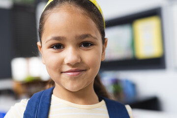 In school, in the classroom, a young biracial girl wearing a backpack is standing