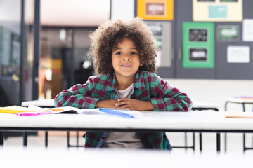In school, young biracial boy leaning on a desk in a classroom, smiling - Powered by Adobe