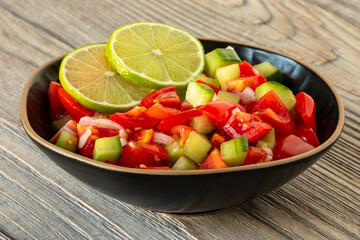 Mexican Pico de Gallo salsa with cucumber in a bowl. Two lime slices. Rustic wooden table.