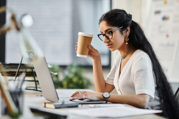 A indian woman, coffee in hand, sits at a desk in a serene moment of contemplation.