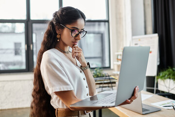 Young indian woman with glasses working on laptop in office setting.