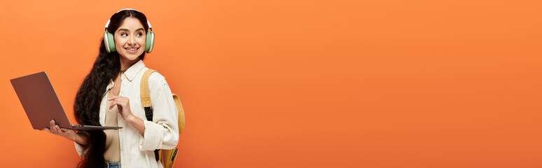 Young indian woman holds laptop while striking a pose in front of wall.