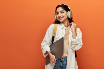 Young indian woman with headphones, holding laptop on orange background.