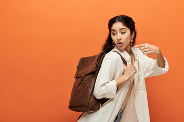 A young indian woman with a brown backpack poses against a vivid orange background.