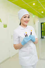 Vertical portrait of smiling female nurse holding test tubes with blood standing on hallway in...