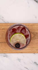 Cocktails. Top view of a Strawberry Jägerinha, with ice, lime and a blueberry, on a wooden tablet on the white marble table with a wooden background.	
