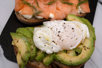 Healthy cuisine. Gourmet sandwich. Top view of a smoked salmon, cream cheese, sliced avocado and poached eggs sandwich with fried potatoes on a black dish on the white marble table.	