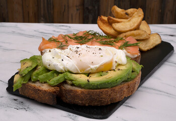 Healthy cuisine. Gourmet sandwich. Top view of a smoked salmon, cream cheese, sliced avocado and poached eggs sandwich with fried potatoes on a black dish on the white marble table.	