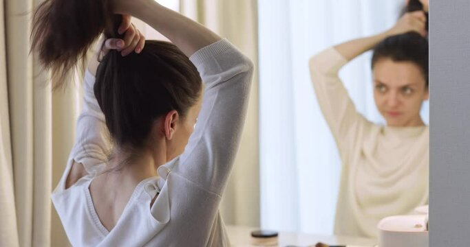 woman puts her long hair in bun and looks at mirror in bedroom