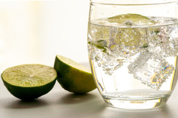 A glass of sparkling water and a slice of lime on ice with a light background. Two lime halves next to the fresh, chilled drink