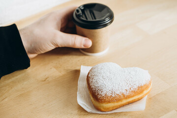 A paper cup of coffee and a heart-shaped donut in male hands.