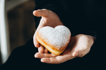 A man in a black hoodie holds a heart-shaped donut covered in powdered sugar.