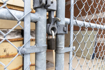 Close-up view of sturdy metallic padlock - chain-link fence gate - rusted metal barrels in...