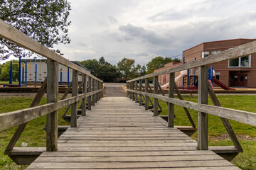 Obraz premium Wooden bridge leading to playground and school building - surrounded by green grass and trees under a cloudy sky. Taken in Toronto, Canada.