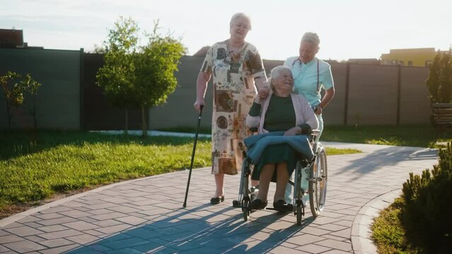 A geriatric nurse takes care of pensioners in front of a nursing home, takes them for a walk in the fresh air, pushing an old woman in a wheelchair