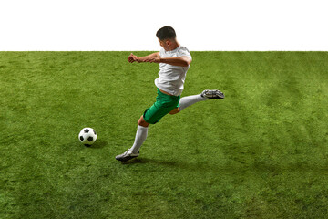 Action shot of young athletic guy, footballer in green and white gear, executing powerful kick on grassy pitch isolated white backdrop. Concept of professionals sport, competition, tournament, action.