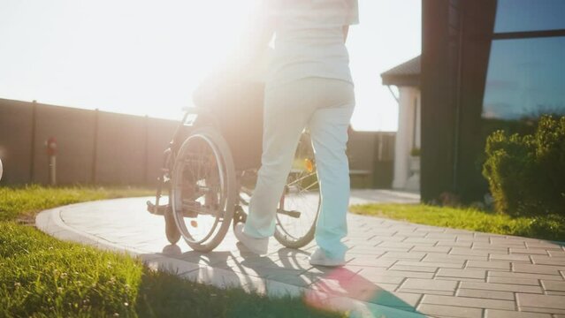 A nurse pushes an old woman in a wheelchair along a path in the yard in front of a nursing home