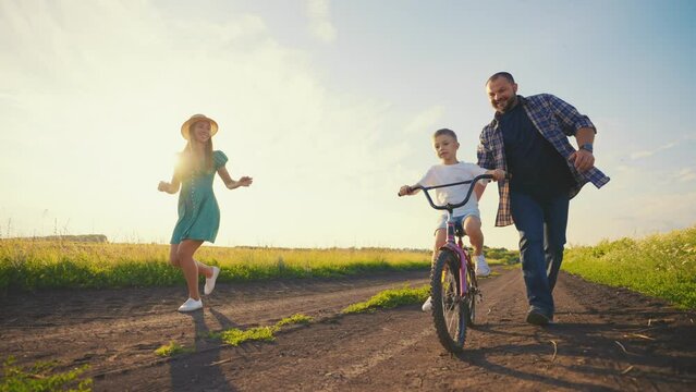 Childhood bike concept. Mother and dad teaching son to ride bicycle. Young family teaching son to ride bike first time on countryside rural road at sunset. Happy kid concept. The best moments together