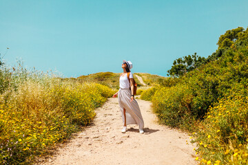 A young girl is walking along the path with a backpack and wearing a white dress. girl in a dress and with a backpack. Happy and freedom-loving woman enjoying a walk.