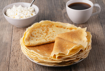 A stack of thin pancakes, a plate with cottage cheese and coffee on a wooden table