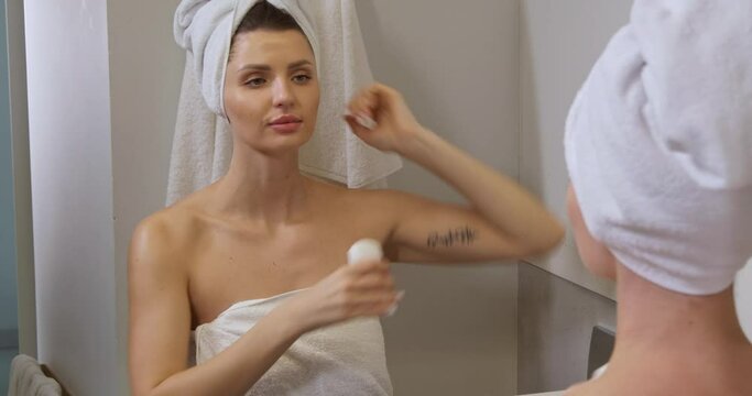 Beautiful woman with wet hair wrapped in towel applying solid deodorant near mirror in bathroom