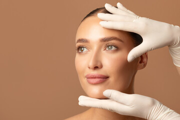 Cosmetic surgery concept. Doctor examining young woman's face, beautiful lady during visit to cosmetologist cabinet, beige studio background, copy space