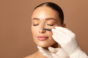 Rhinoplasty concept. Doctor's gloved hand making marks on female face, young woman posing on beige...