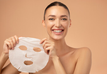 Beauty portrait of young European woman holding facial sheet mask looking and smiling at camera...