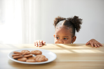 African child girl hiding and looking chocolate cookies or biscuits on dish from under the table