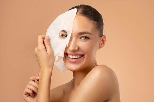 Young pretty European woman removing moisturizing, rejuvenating white fabric sheet mask from her face after using it, smiling at camera, beige background