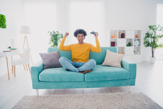 Full size photo of nice young man hold controller raise fist wear yellow pullover white interior apartment indoors