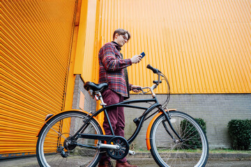 Young hipster man using mobile phone while standing near bicycle near orange wall. Student using 5g...