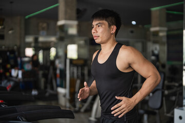 A determined man in a black tank top runs on a treadmill in a modern gym. His muscular build and...