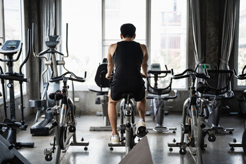 A man in a black tank top rides a stationary bike in a modern gym, surrounded by other exercise...