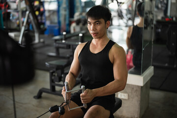 A man in a black tank top works out on a seated row machine in a modern gym. His serious expression...