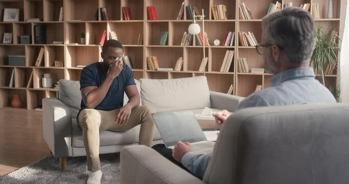 African American male sitting on soft grey coach and beginning crying. Psychotherapist sitting in front of him and taking notes. Listening carefully to man and thinking about best solution.