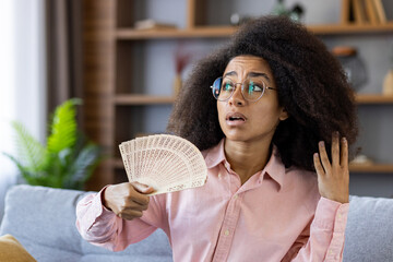 A woman with glasses cools down with a fan in a living room. She looks tired, sitting on a couch...