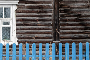 White window on the wall of a log house. A blue fence in front of a wall of a log house. Old logs...