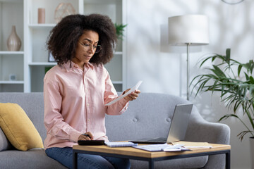 An organized woman manages household finances with a laptop and a calculator in her modern home...