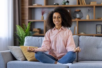 A woman with curly hair and glasses is meditating on a gray couch in a serene living room, smiling...