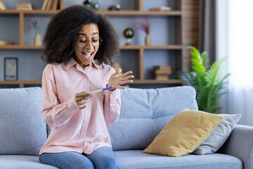 A woman in a cozy living room joyfully looks at a positive pregnancy test, anticipating future...