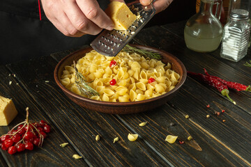 The chef adds hard cheese to a plate of pasta using a grater. Working environment on the kitchen table in the cook room. Delicious pasta with vegetables and aromatic spices