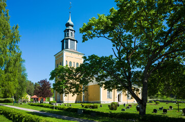 Yellow and white church building surrounded by a graveyard green lawn with gravestones under a large spreading tree under a blue sky, Ramnäs church in Sweden