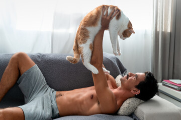 young man lying on a sofa plays with a brown and white cat