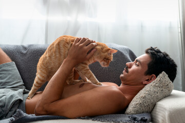 young man lying on a sofa  interacts with a brown tabby cat  