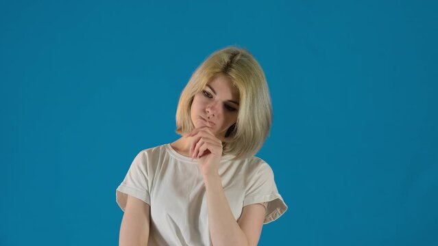 Young woman student bites lip thinking about future exam on blue background. Thoughtful blonde female touches chin slow motion