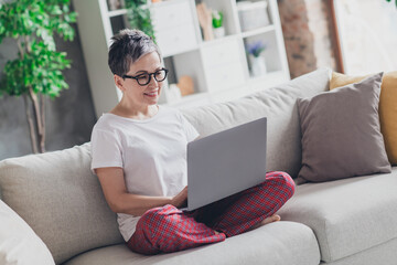 Full size photo of nice senior woman work netbook sit sofa dressed white clothes cozy living room...