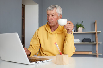 Confident senior man wearing yellow hoodie using laptop at home in living room sending email and working at home freelancer typing on computer and drinking coffee